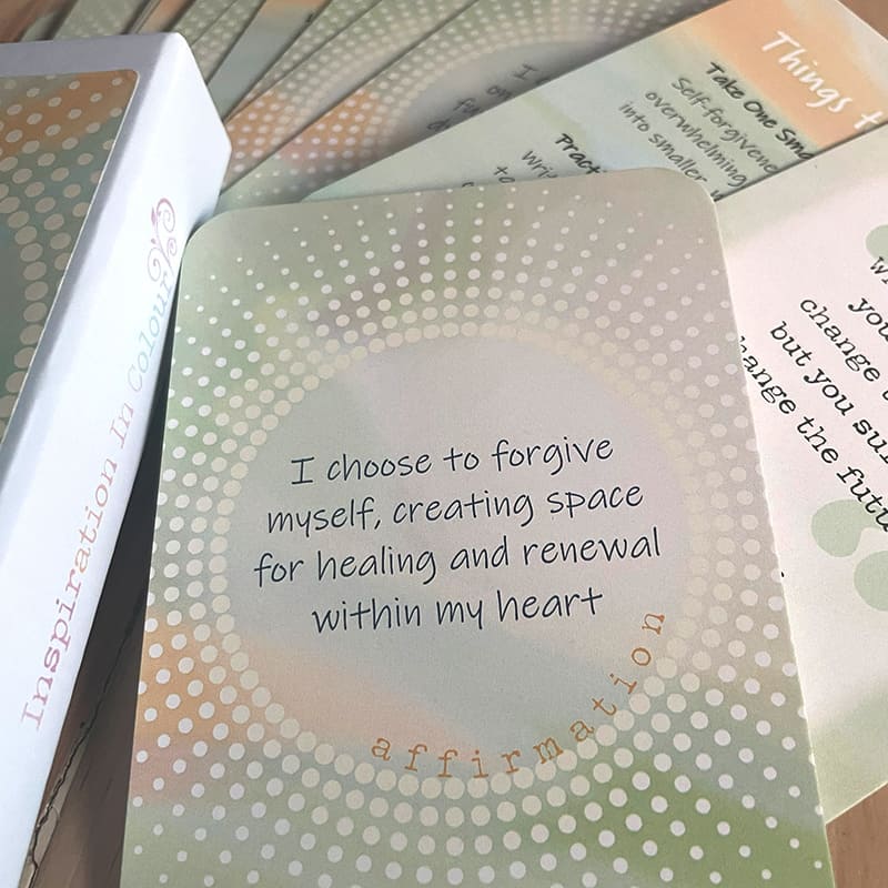 A close-up of a card reading "I choose to forgive myself, creating space for healing and renewal within my heart – affirmation," beside the closed card box labeled "Inspiration In Colour," with other cards fanned behind it.