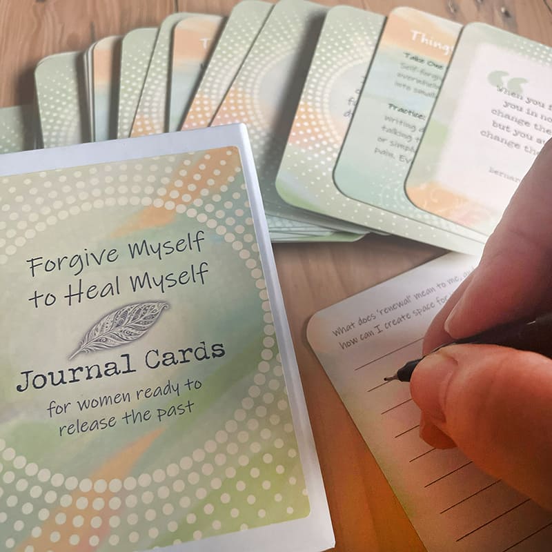 The closed "Forgive Myself to Heal Myself Journal Cards" box next to a hand writing on a lined journal card prompting "What does 'renewal' mean to me, and how can I create space for it?", with fanned prompt and quote cards above.