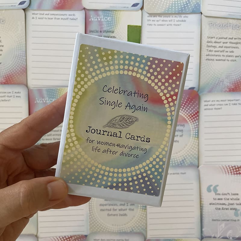 A hand holds the closed "Celebrating Single Again Journal Cards" box against a background of scattered cards showing advice text such as "Build a Strong Support System" and a quote reading "You don't have to see the whole staircase, just take the first step.
